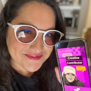 Erica Carrasco, aka Achy Smile, holding a phone showing a photo of her Creative Contributor award graphic from Social Health Awards by Health Union. Migraine awareness wins!