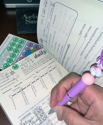 3-Month Physical Symptoms & Mental Health Tracker laying open on a desk with a hand holding a purple butterfly awareness pen writing on an inside page.