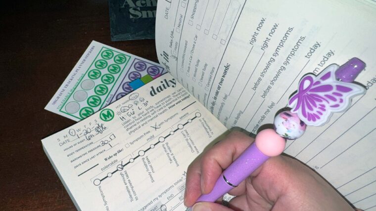 3-Month Physical Symptoms & Mental Health Tracker laying open on a desk with a hand holding a purple butterfly awareness pen writing on an inside page.